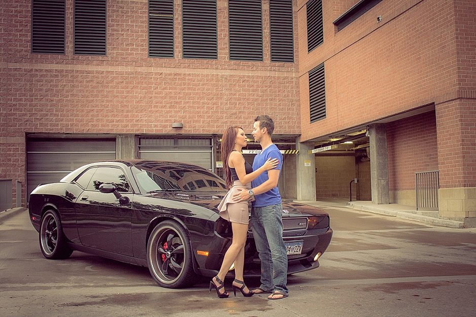 beautiful engaged couple in front of a black dodge challenger